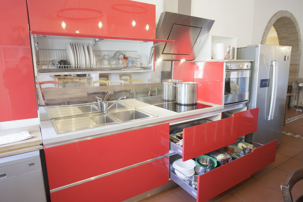 A modern kitchen features vibrant red cabinetry with sleek lines and stainless steel accents. Two spacious sinks are positioned next to a large induction cooktop. Open drawers showcase a variety of pots and pans, while stacked dishes are visible on shelves above.