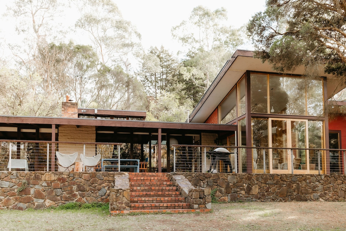 A modern mud-brick home is surrounded by native trees, featuring a spacious covered patio and large glass windows. The entrance includes stone steps leading to a well-defined outdoor area, where outdoor furniture is positioned for relaxation.