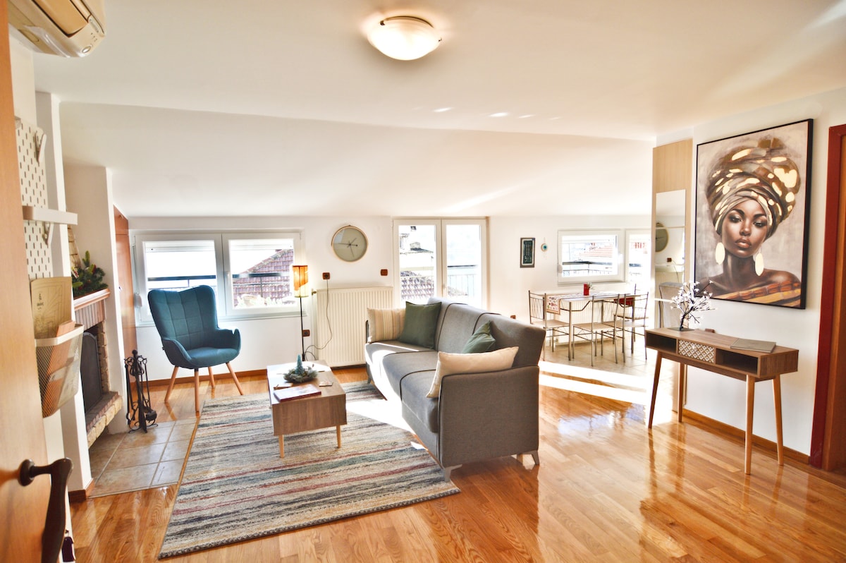 A bright living space features large windows allowing natural light to fill the room. Comfortable seating includes a blue armchair and a grey sofa. A wooden coffee table is set on a striped rug, while a dining area with a round table and chairs is visible in the background.