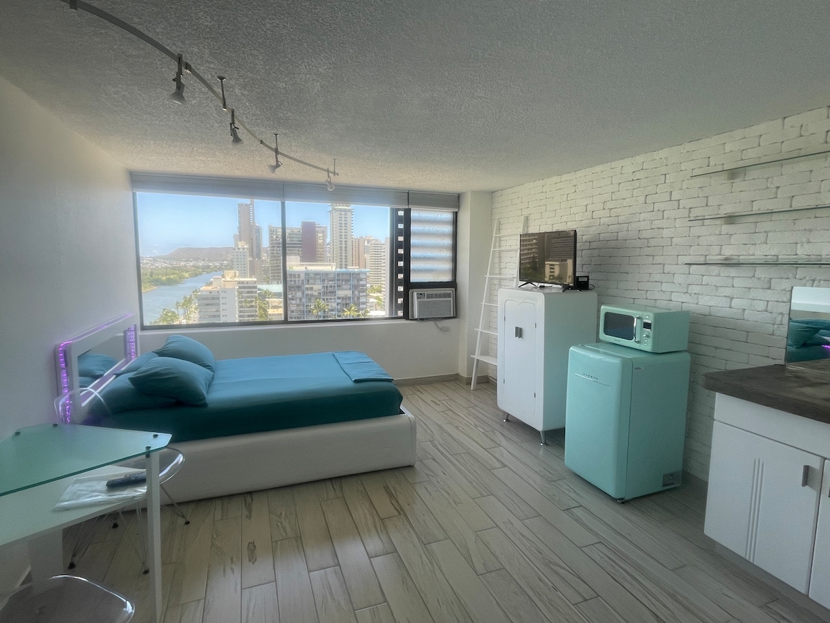 A modern studio features a spacious bed with teal bedding, situated beside a large window offering city and ocean views. A glass desk and chair are positioned to the left, while a small kitchenette with retro appliances is visible in the corner.