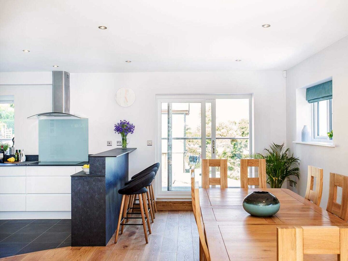 An open-plan kitchen and dining area features sleek white cabinetry and a dark countertop. A wooden dining table with stylish chairs is positioned in natural light. Glass doors lead to an outdoor balcony, with vibrant greenery visible in the background.