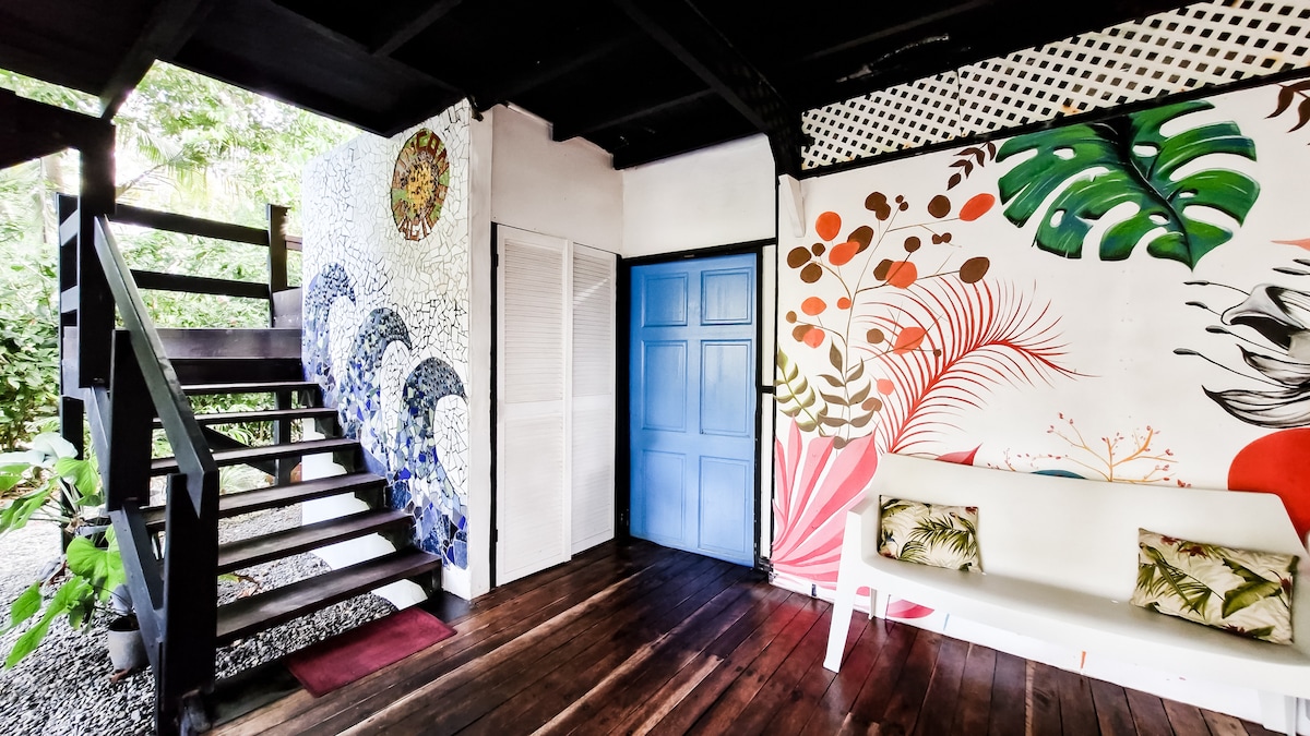 A vibrant entrance area features colorful tropical wall art, enhancing the inviting atmosphere. A light-colored sofa is positioned against the wall, accompanied by decorative cushions. Stairs lead to the upper level, while natural light illuminates the wooden flooring and nearby greenery through open spaces.