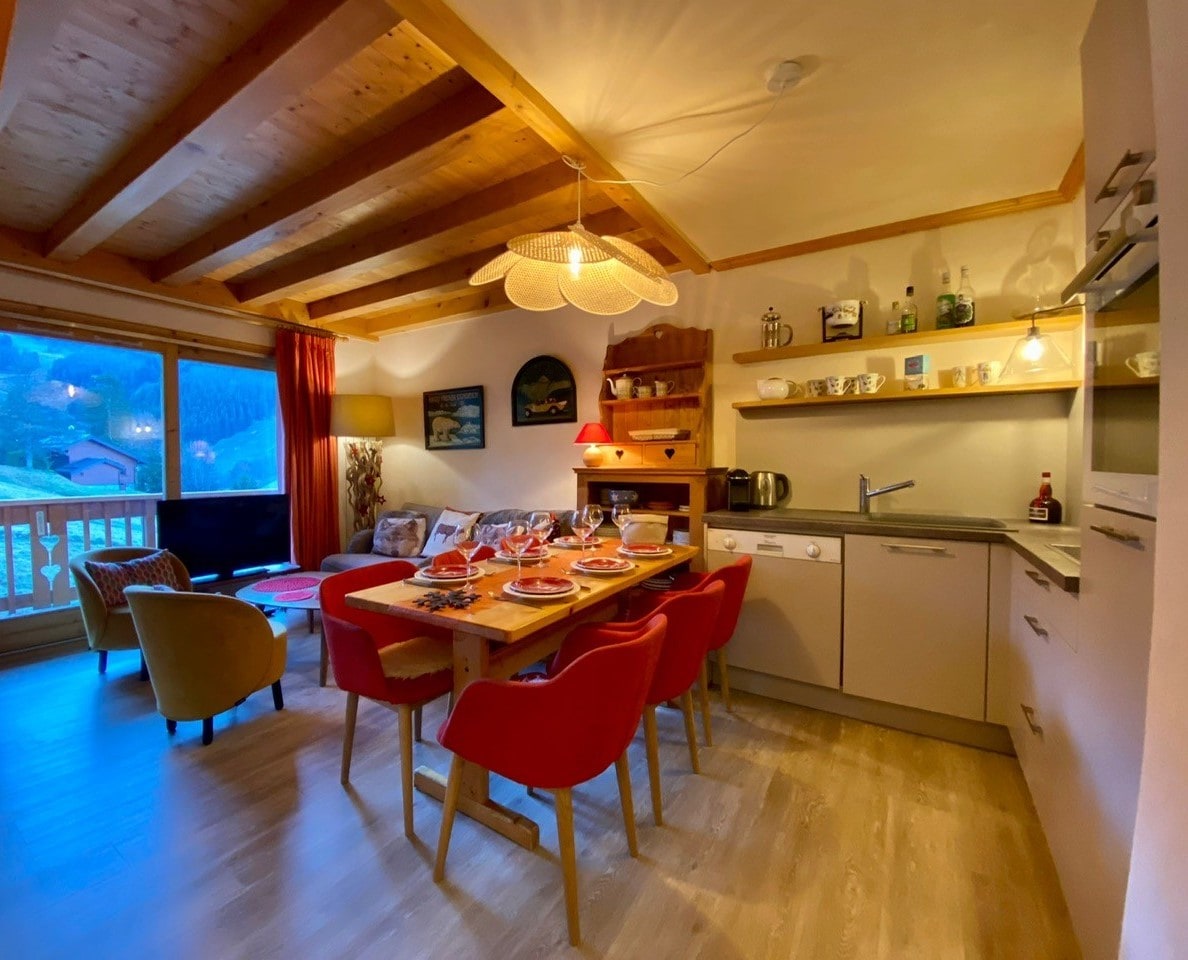 A welcoming dining area is complemented by a wooden ceiling and large windows offering views of the surrounding landscape. A wooden table is set for dining, surrounded by colorful chairs. Modern appliances are visible in the kitchen space, and decor elements add a homey touch.