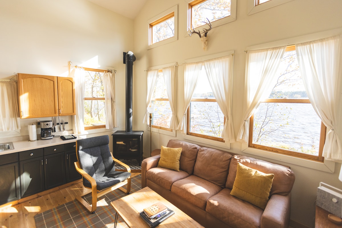A spacious living area features large south-facing windows that fill the room with natural light and provide views of the lake. A comfortable sofa is paired with a wooden chair, and a coffee table sits in the center. The kitchen is equipped with wooden cabinetry and essential appliances.