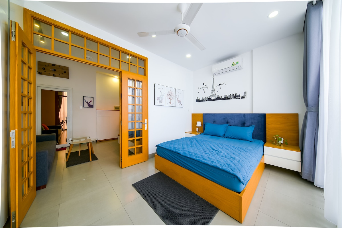 A modern bedroom features a spacious blue bed with a matching bedspread. Natural light enters through large windows framed by soft curtains. The room includes a wooden bedside table and bright orange doors, leading to a small living area visible in the background.