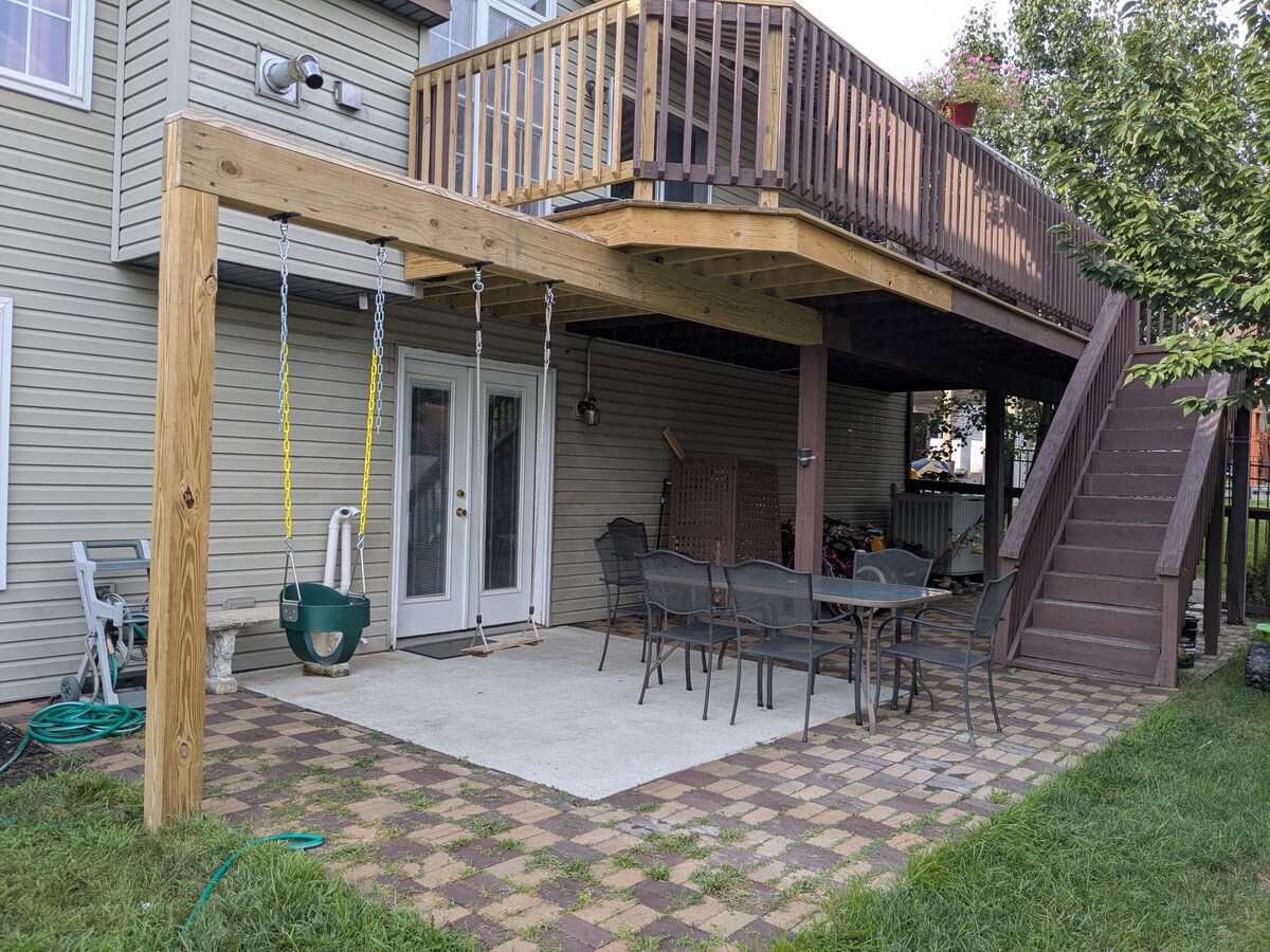 A covered outdoor seating area features a patio with a set of rectangular tables and chairs, suitable for dining or relaxation. Above, a wooden deck provides additional space, supported by sturdy beams. A swing and grassy area offer options for leisure and play.