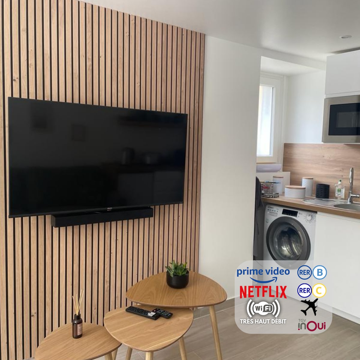 A modern living area features a flat-screen television mounted on a wooden accent wall. A small round table with two chairs is positioned nearby. The kitchen area is visible, showcasing a washing machine and various kitchen appliances.
