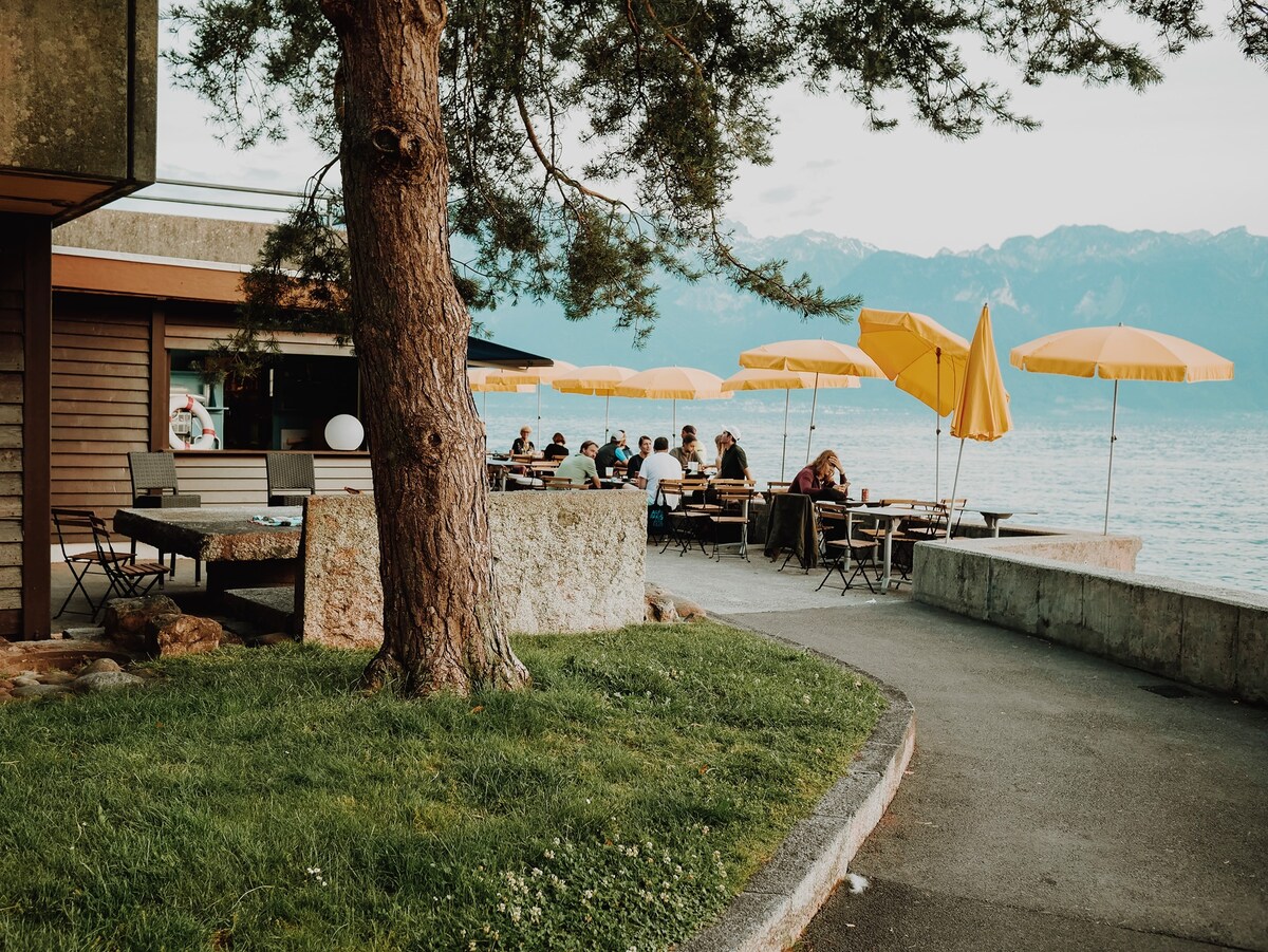 A waterfront café is depicted with several outdoor tables shaded by large yellow umbrellas. Guests are seated at the tables, enjoying the tranquil view of the lake and surrounding mountains. The pathway is lined with greenery, and a large tree provides natural beauty to the setting.