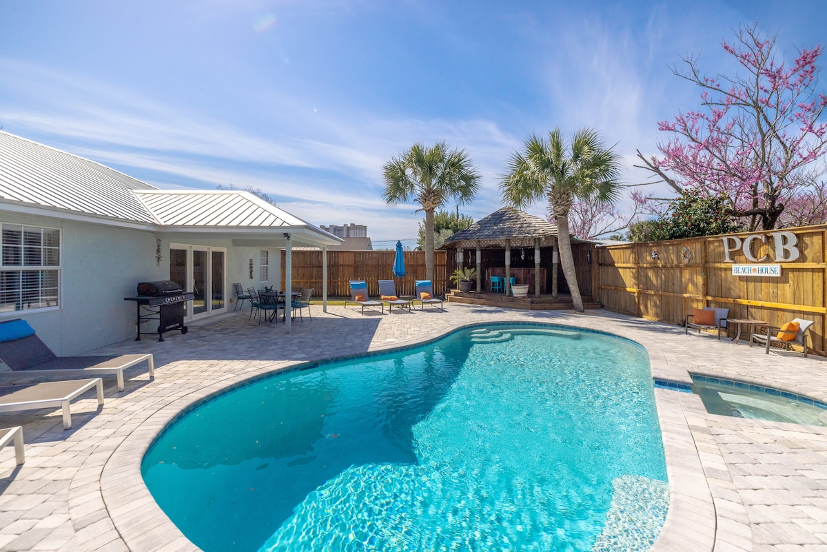The outdoor area features a refreshing private pool surrounded by a spacious stone patio. Lounge chairs are positioned around the pool, and a shaded Tiki bar is visible in the background, framed by palm trees and a wooden fence.