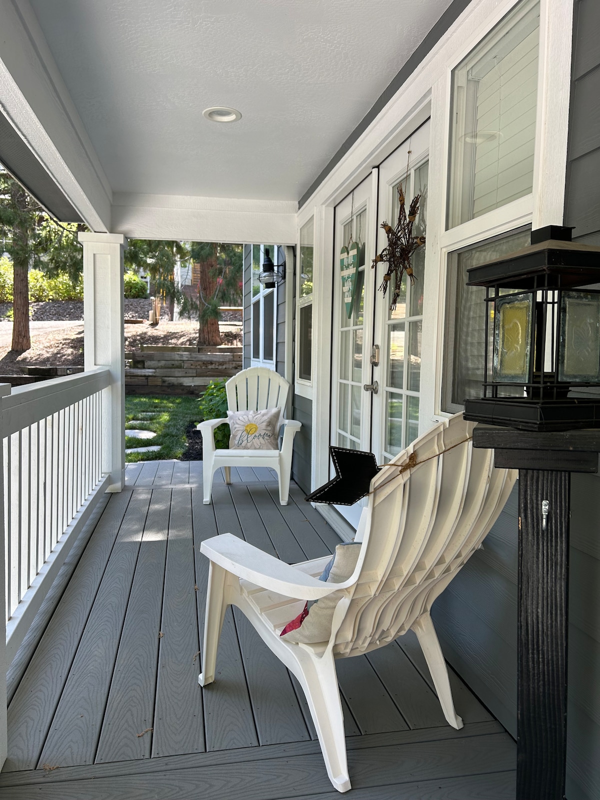 A welcoming porch area features two white chairs placed beside a small side table. Sunlight filters through the glass doors, illuminating the space. A decorative lantern stands nearby, adding a touch of charm, while a small cushion brings comfort to one chair. Lush greenery is visible beyond.