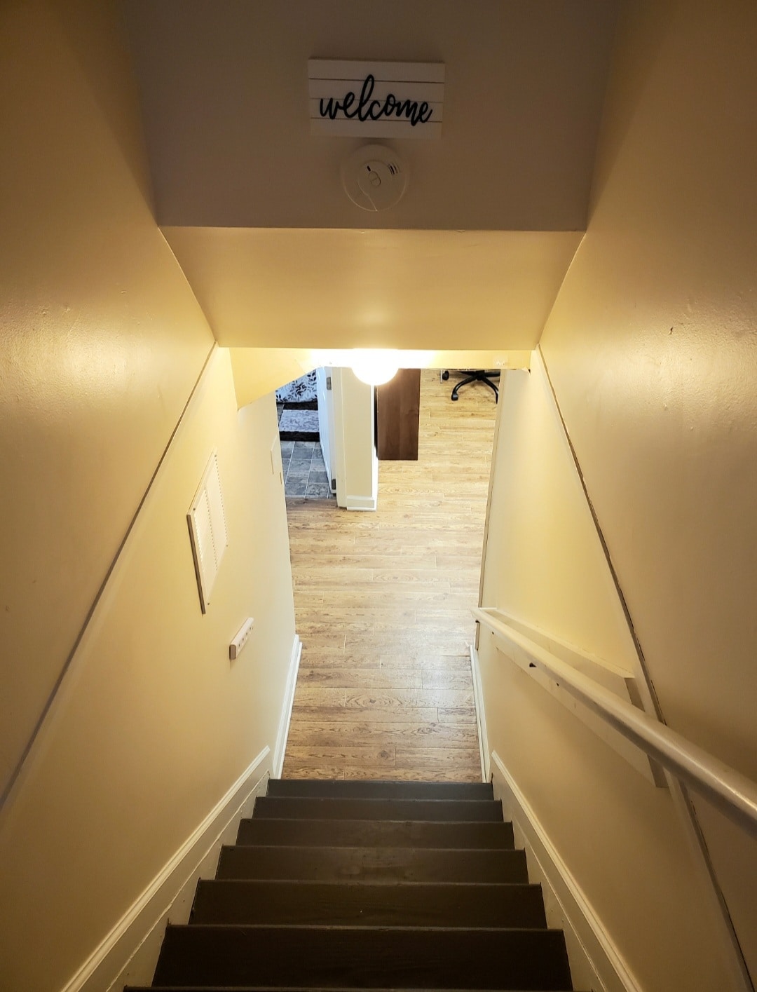 A stairway leads down to the basement apartment, featuring light-colored walls and a light fixture illuminating the steps. A wooden welcome sign is positioned at the top of the stairs, adding a homey touch. The space is designed for easy access to the living area below.