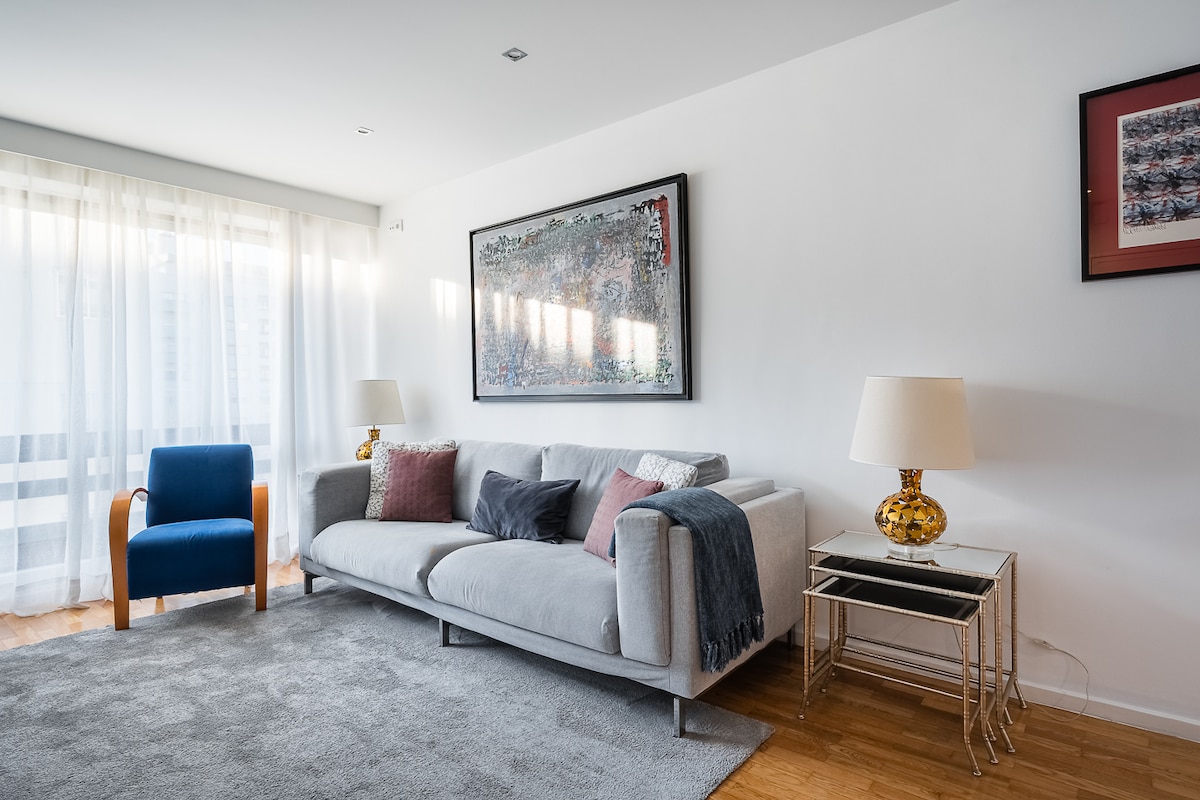 A modern living area is displayed, featuring a light grey sofa adorned with assorted pillows, a blue chair, and a stylish table with a decorative lamp. Large windows allow natural light to fill the space, complemented by sheer white curtains.