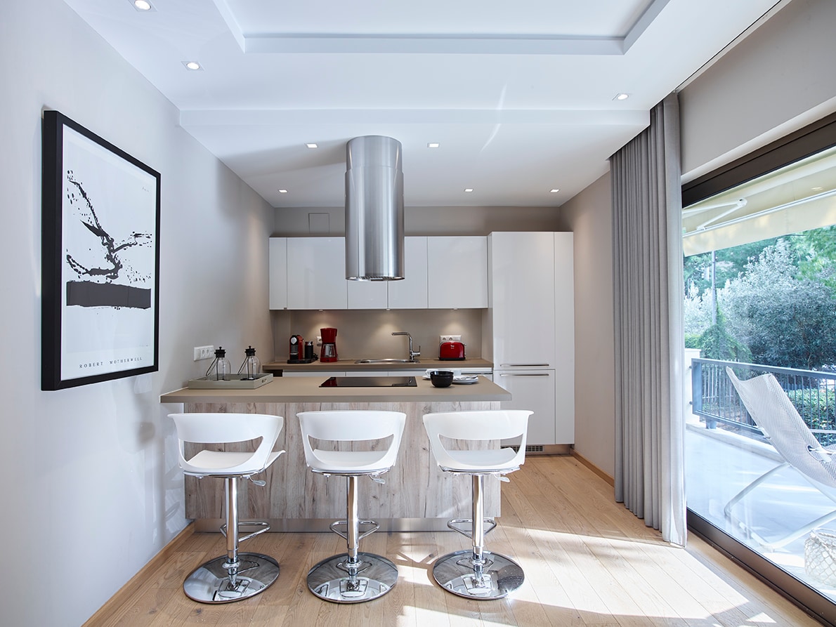 A modern kitchen area is featured with sleek white cabinetry and stainless steel appliances. Three bar stools are arranged at the island, which faces large glass doors that open to a patio area. Warm wooden flooring complements the neutral tones of the walls and furnishings.