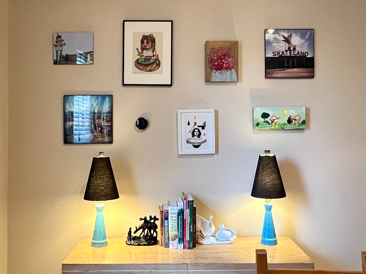 A gallery wall displays a variety of framed artwork and photographs above a wooden console table. Two decorative lamps with black shades provide ambient lighting, while a collection of books and art objects is arranged on the table, enhancing the room's creative atmosphere.