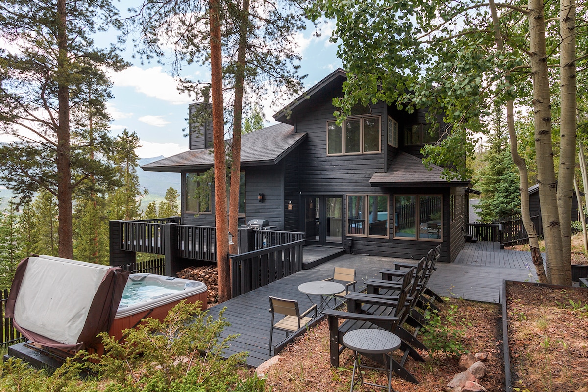 A modern mountain retreat is surrounded by trees, showcasing an expansive outdoor deck with ample seating. A state-of-the-art hot tub is positioned nearby, providing access to serene views. Large windows of the home reflect the natural surroundings, creating a harmonious connection with the landscape.