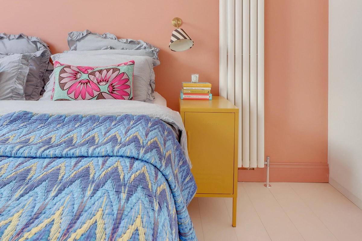 A bedroom features a spacious bed with a patterned quilt in shades of blue and yellow. A colorful decorative pillow rests against the headboard. A vibrant nightstand in yellow holds a stack of books, and a wall-mounted lamp illuminates the space. The wall is painted a soft coral.
