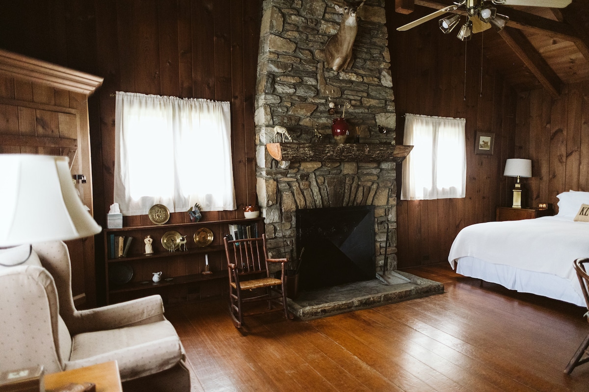 A cozy interior space features a stone fireplace at the center, adorned with a mounted deer trophy. Soft curtains frame two windows, allowing natural light to illuminate the room. An inviting queen-sized bed and rustic furniture, including a rocking chair and an armchair, complete the warm atmosphere.