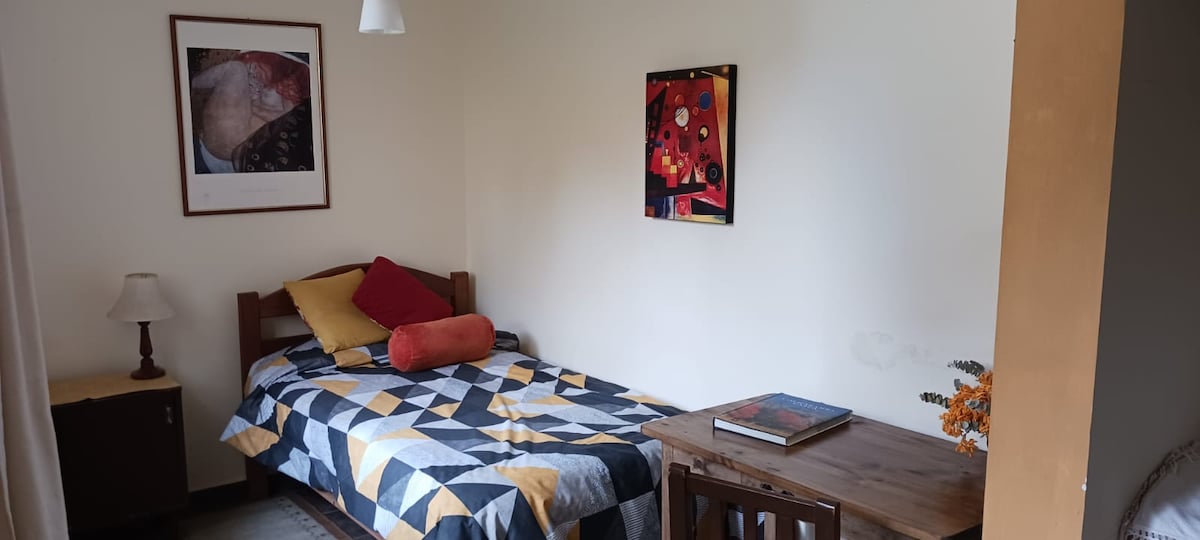 A brightly lit room features a spacious bed adorned with a geometric-patterned blanket and colorful pillows. A wooden desk is positioned nearby, complemented by an artwork on the wall. A lamp and a nightstand add functionality to the inviting space.