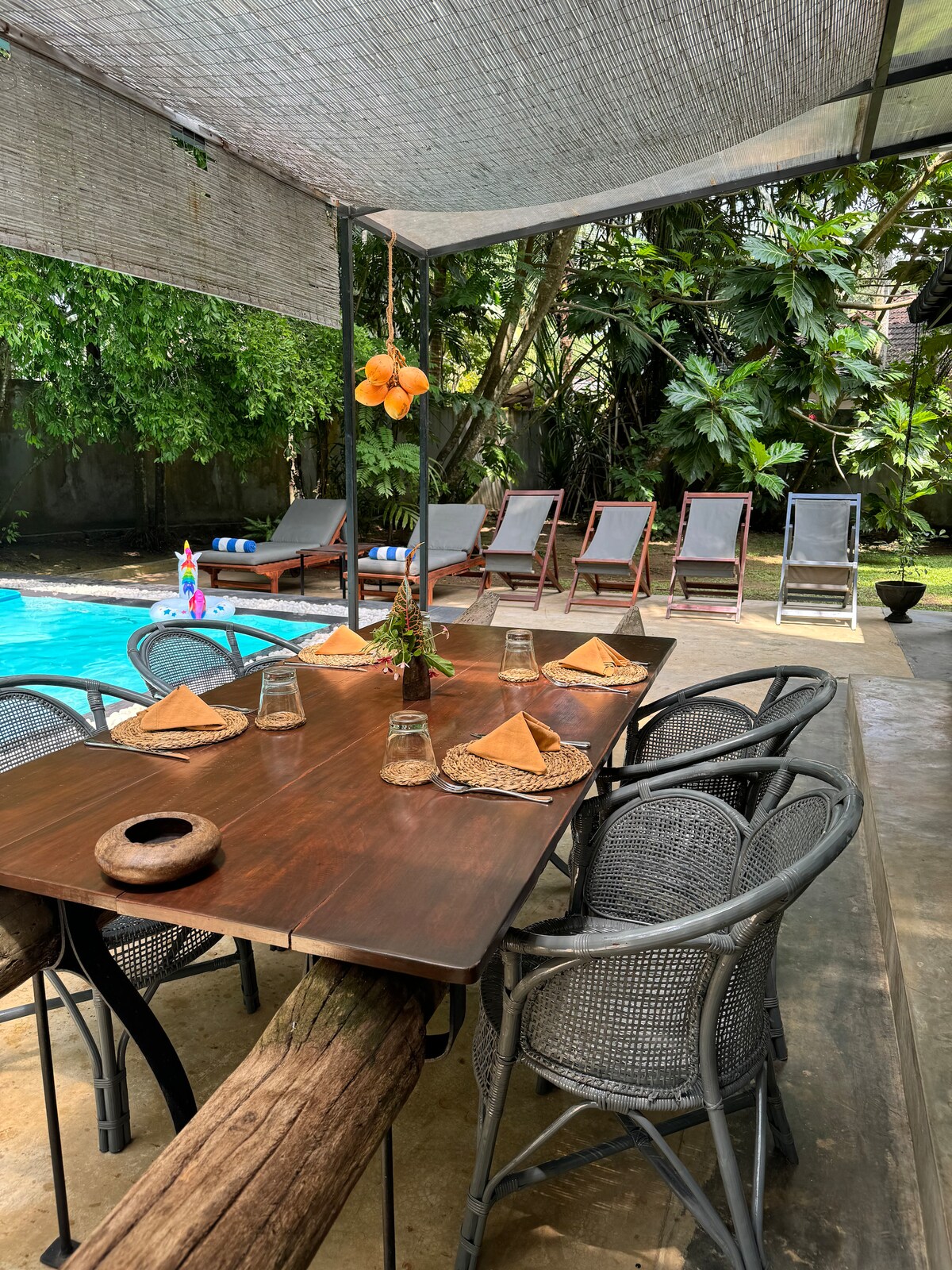 An outdoor dining area is situated under a shade structure, featuring a large wooden table set with woven placemats and decorative napkin folds. Six woven chairs surround the table, while sun loungers line the poolside amidst lush tropical greenery.