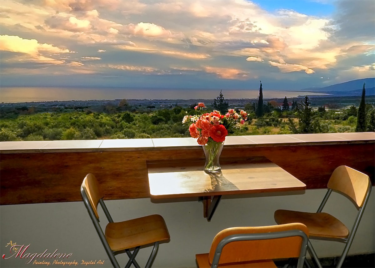 A small outdoor table is set with two wooden chairs on a covered balcony. A vase of colorful flowers is placed in the center of the table. In the background, a panoramic view of the sea and sky, with soft clouds extending over the horizon, is visible.
