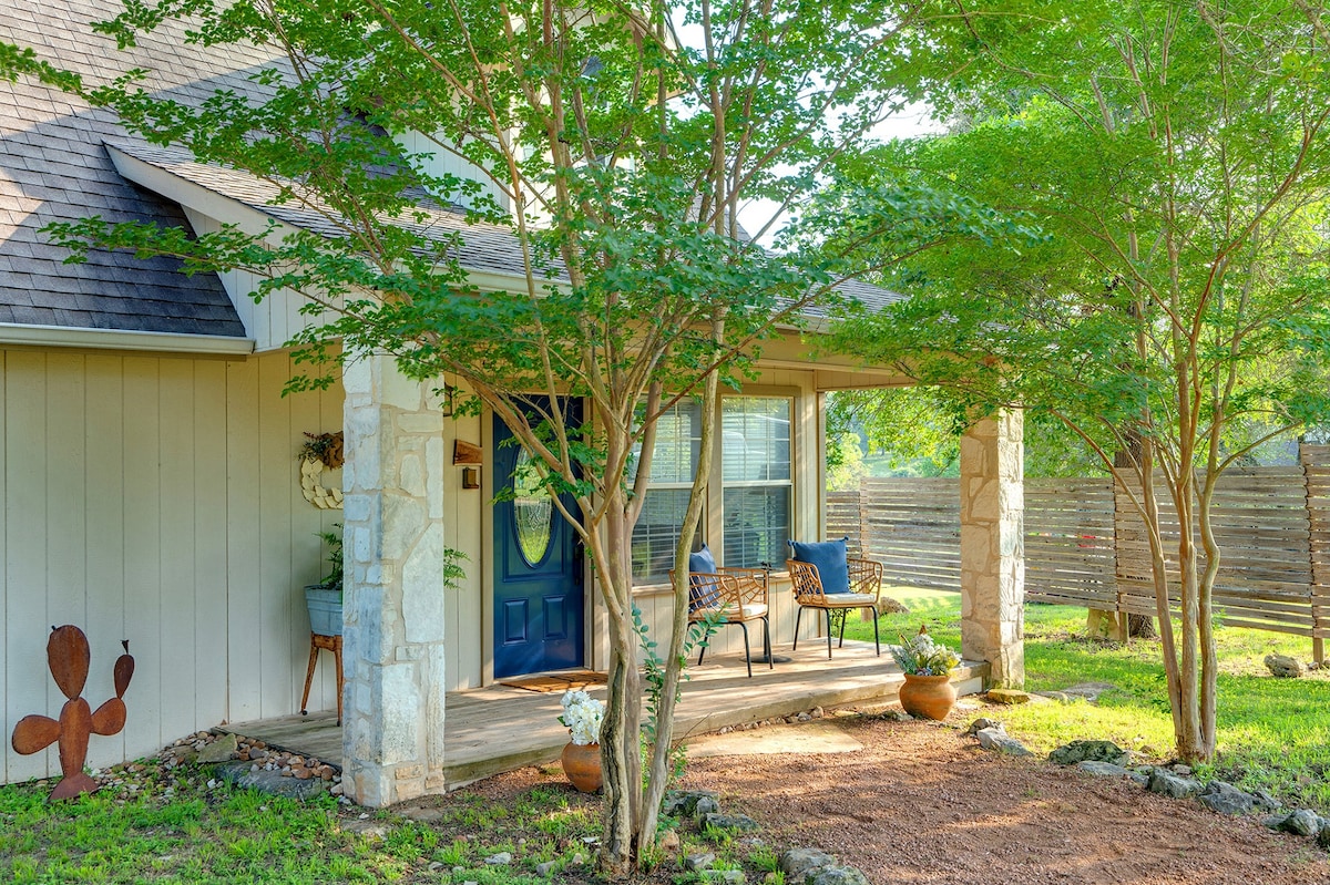 The entrance features a welcoming porch adorned with two chairs and a small table, shaded by trees. Flower pots are placed on the ground, and a stone pathway leads to the door, creating an inviting outdoor space with a natural feel.