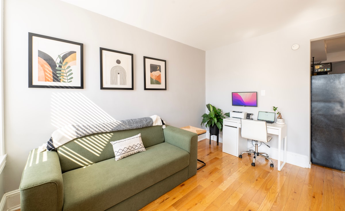 A spacious living area features a green couch complemented by a patterned throw pillow. To the right, a workstation with a monitor and laptop is positioned under ambient light from a nearby window. Art prints adorn the walls, creating a calm atmosphere. Light wood flooring connects the space.