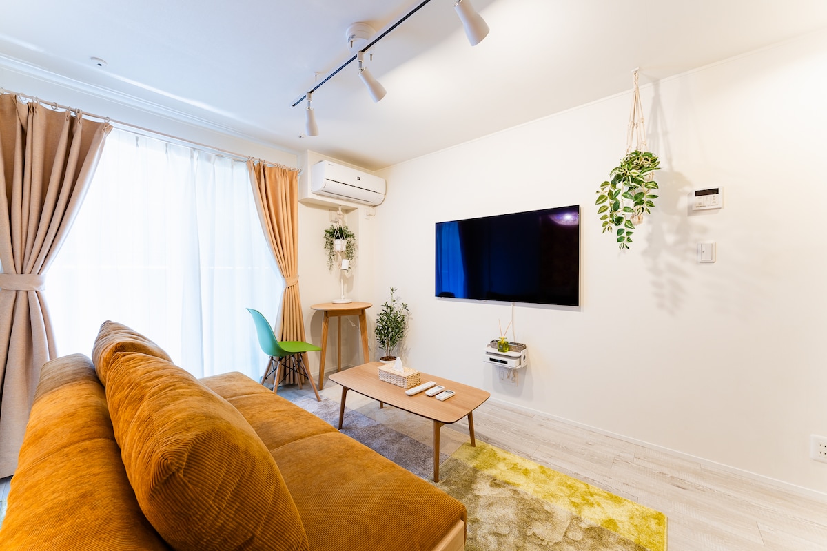 A comfortable living area features a tan sofa facing a wall-mounted television. Natural light filters through sheer curtains, illuminating a small dining table with a green chair adjacent to a decorative plant. A soft, multi-colored rug adds warmth to the space.