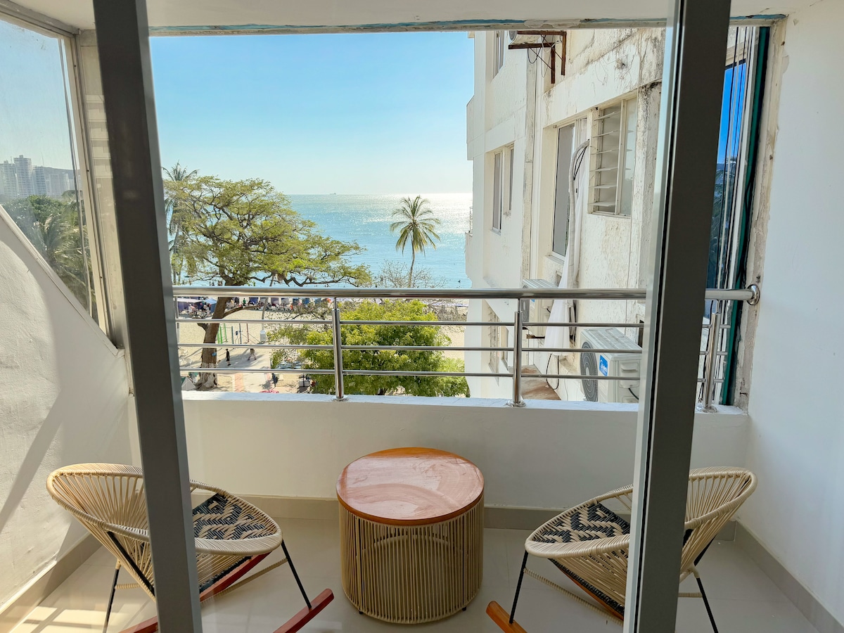 A balcony is seen with two woven chairs and a round table, overlooking a view of the ocean. Bright sunlight reflects off the water, while palm trees sway in the breeze along the beachfront. The railing provides an unobstructed view of the surroundings.