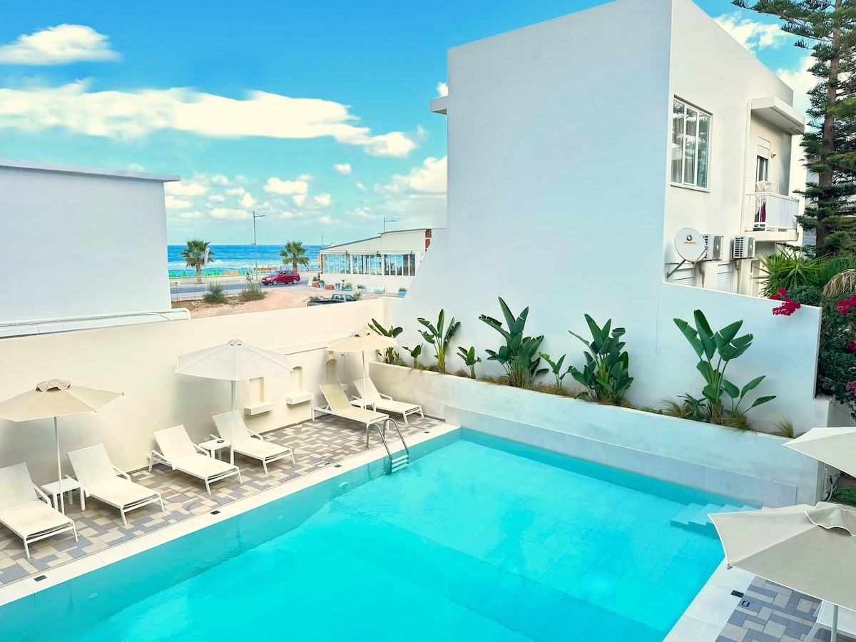 A refreshing pool area is highlighted, featuring a clear blue pool surrounded by sun loungers and umbrellas. Lush greenery borders the space, with a view of the beach and ocean visible in the background under a bright sky.