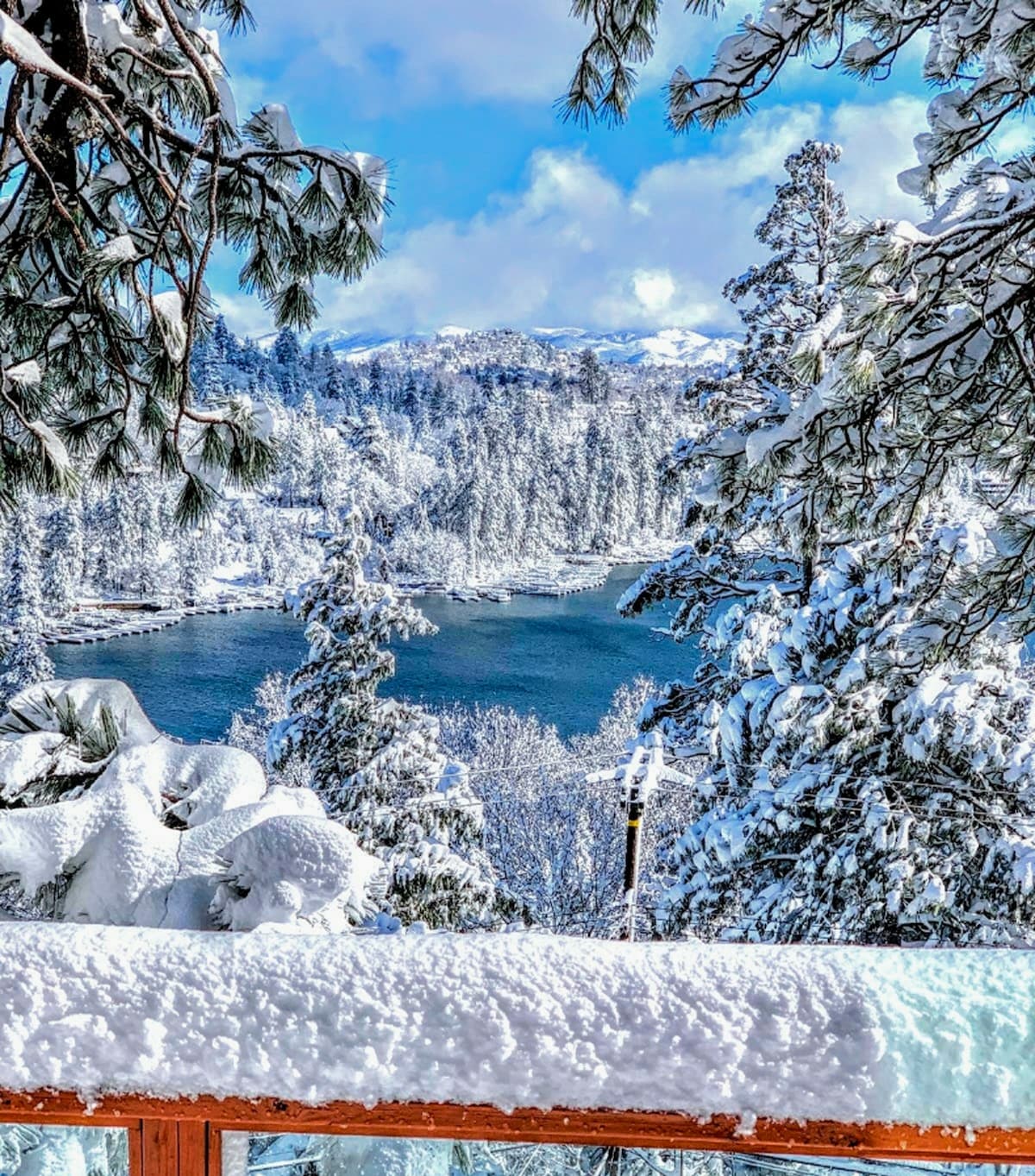 A winter scene showcases a snow-covered landscape with tall, frosty trees framing a serene view of Lake Arrowhead. The reflection of the surrounding snow-laden mountains glimmers on the lake's surface under a bright blue sky, creating a tranquil atmosphere.