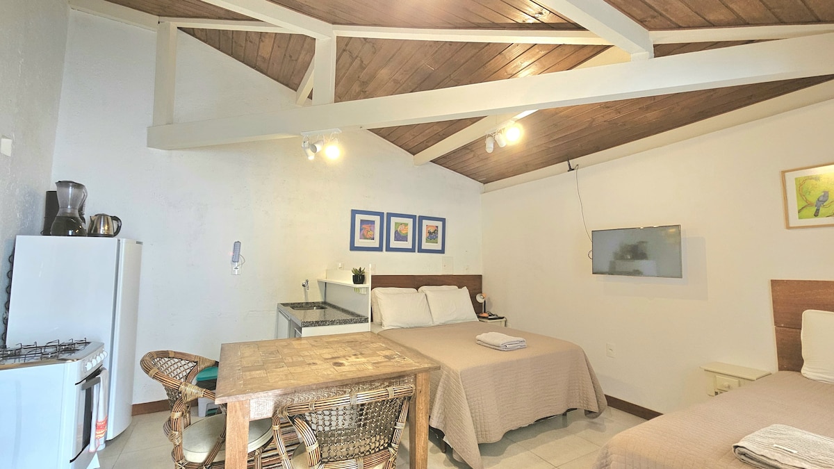 The studio features a spacious layout with a wooden beam ceiling. Two beds are positioned on one side, with a kitchenette and dining area visible across from them. A dining table with chairs complements the room, while a wall-mounted TV provides entertainment.