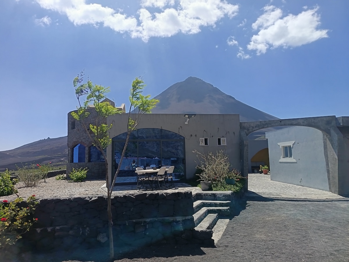 The exterior of the house is visible, showcasing a stone structure with large windows. A dining area with a table and chairs is arranged outside, overlooking a volcanic landscape. The surrounding garden features local flora, while the imposing volcano is seen in the background under a clear blue sky.