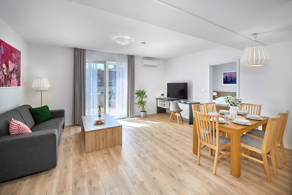 A bright and airy living space features a comfortable gray sofa and a wooden coffee table. A dining area with a wooden table and six chairs is visible. Large windows allow natural light to fill the room, and a television is mounted on the wall.