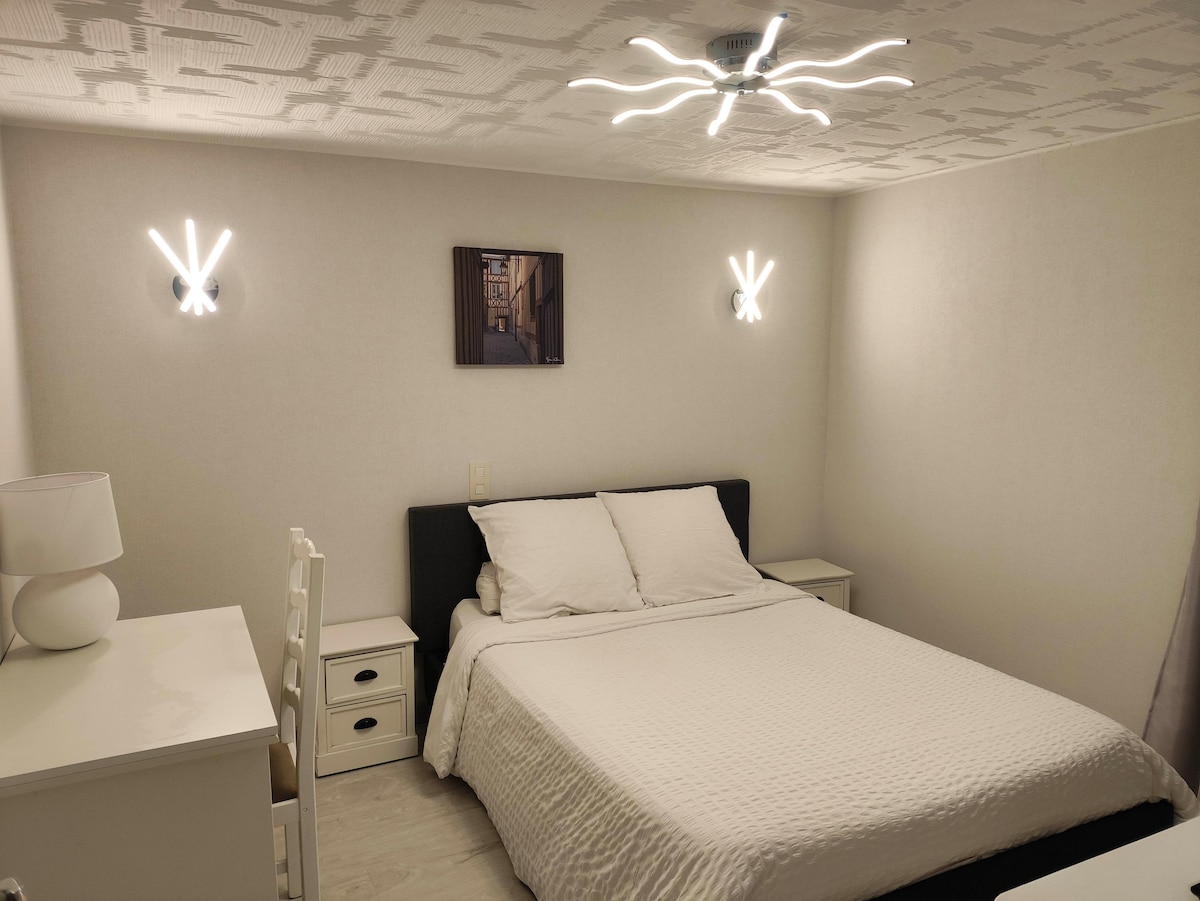 A well-lit bedroom features a spacious bed with a white quilt and two pillows. Modern wall lighting flanks the bed, offering soft illumination. A small bedside table sits beside the bed, and a stylish chair and dresser are present in the room, enhancing functionality.
