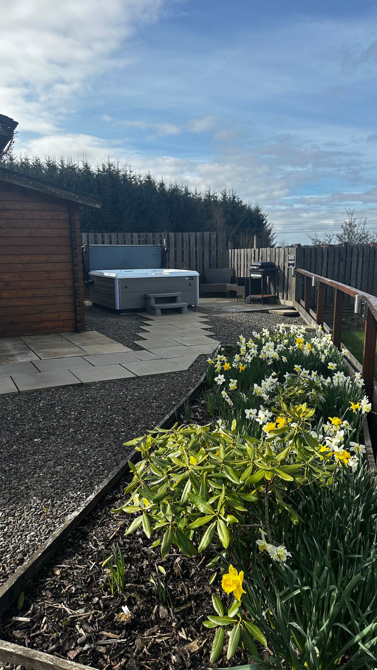 The outdoor area features a private hot tub alongside a spacious wooden cabin. A stone pathway leads through blooming flower beds filled with daffodils. Comfortable seating is arranged nearby, offering a relaxed setting amidst the greenery and natural surroundings.