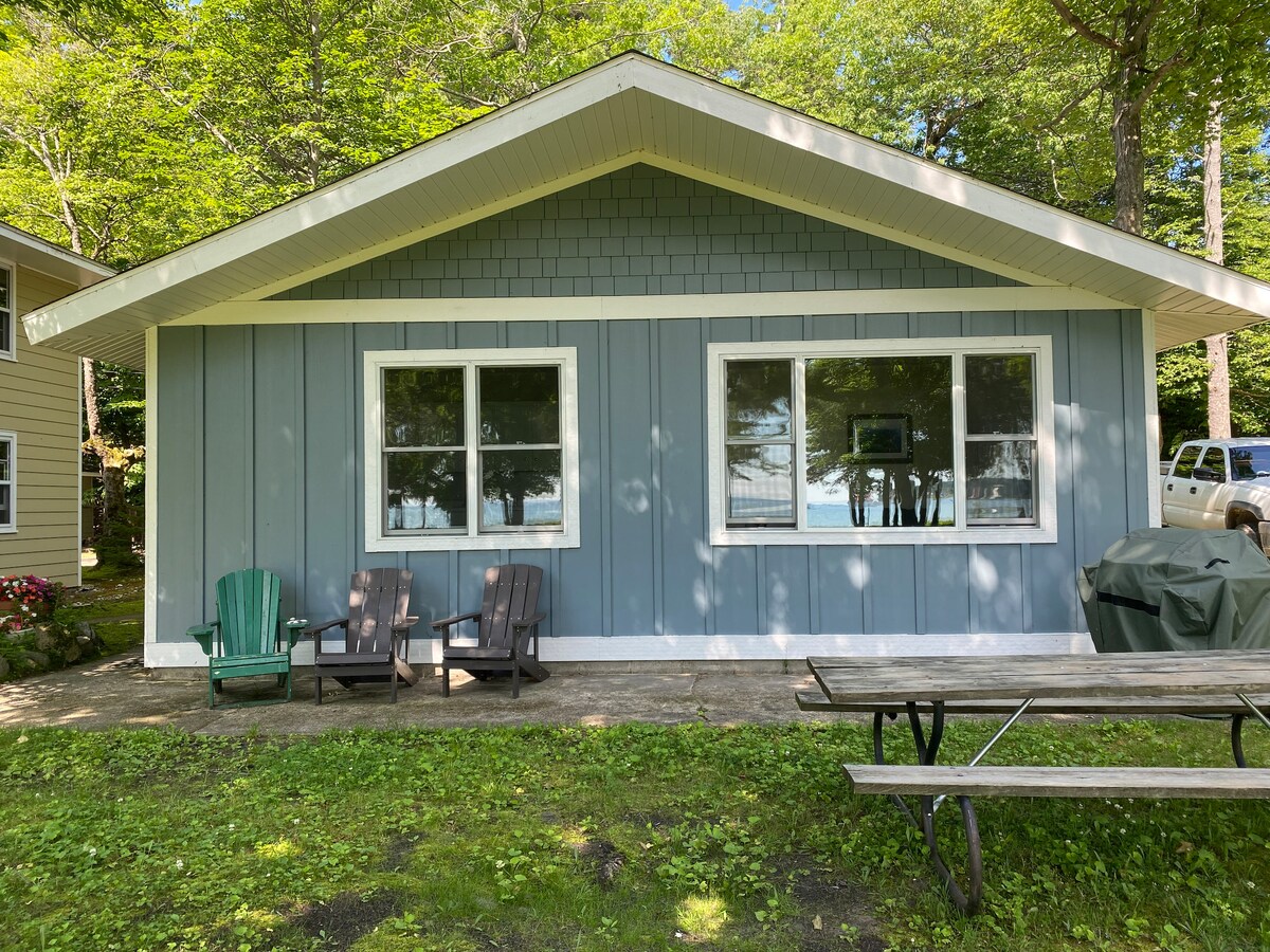 A cottage exterior is shown, featuring two large windows that allow natural light to brighten the interior. Two green chairs are positioned on the patio beside a picnic table. A gas grill is noticed nearby, all set against a backdrop of lush greenery.