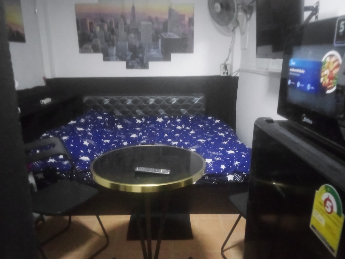 A compact living space showcases a large bed with a starry blue coverlet, complemented by a black leatherette headboard. A small round table with two chairs is positioned near a wall-mounted TV, offering a cozy area for dining or relaxation.