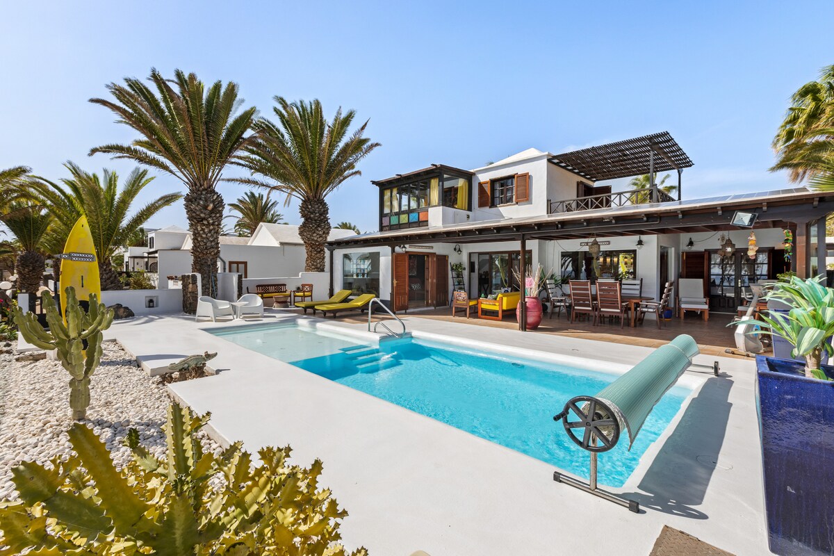 A spacious outdoor area features a private pool surrounded by tropical palm trees. Sun loungers and a seating area are positioned alongside the pool, while a patio with a dining area is visible in the background. The modern villa showcases large windows allowing natural light.