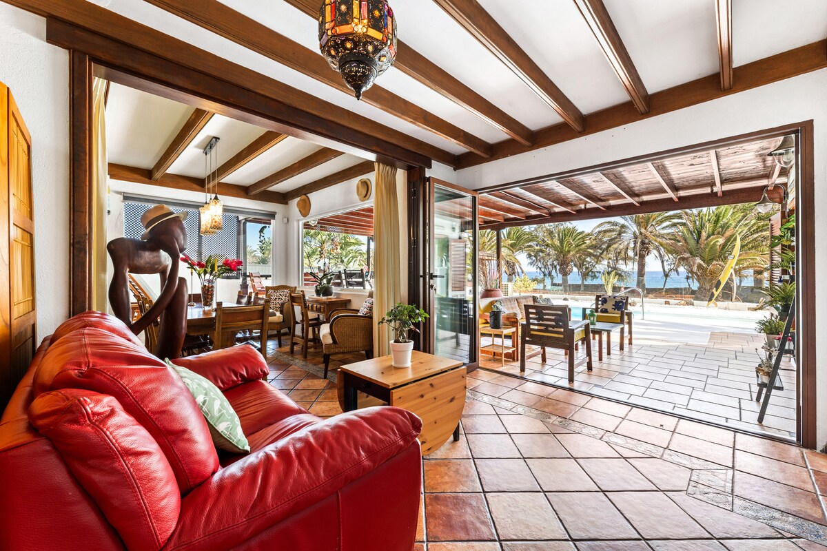 A spacious living area features a red leather sofa and a wooden coffee table, with natural light flowing through large sliding glass doors that open to a patio. The setting includes wooden beams on the ceiling and a dining area with a view of palm trees outside.