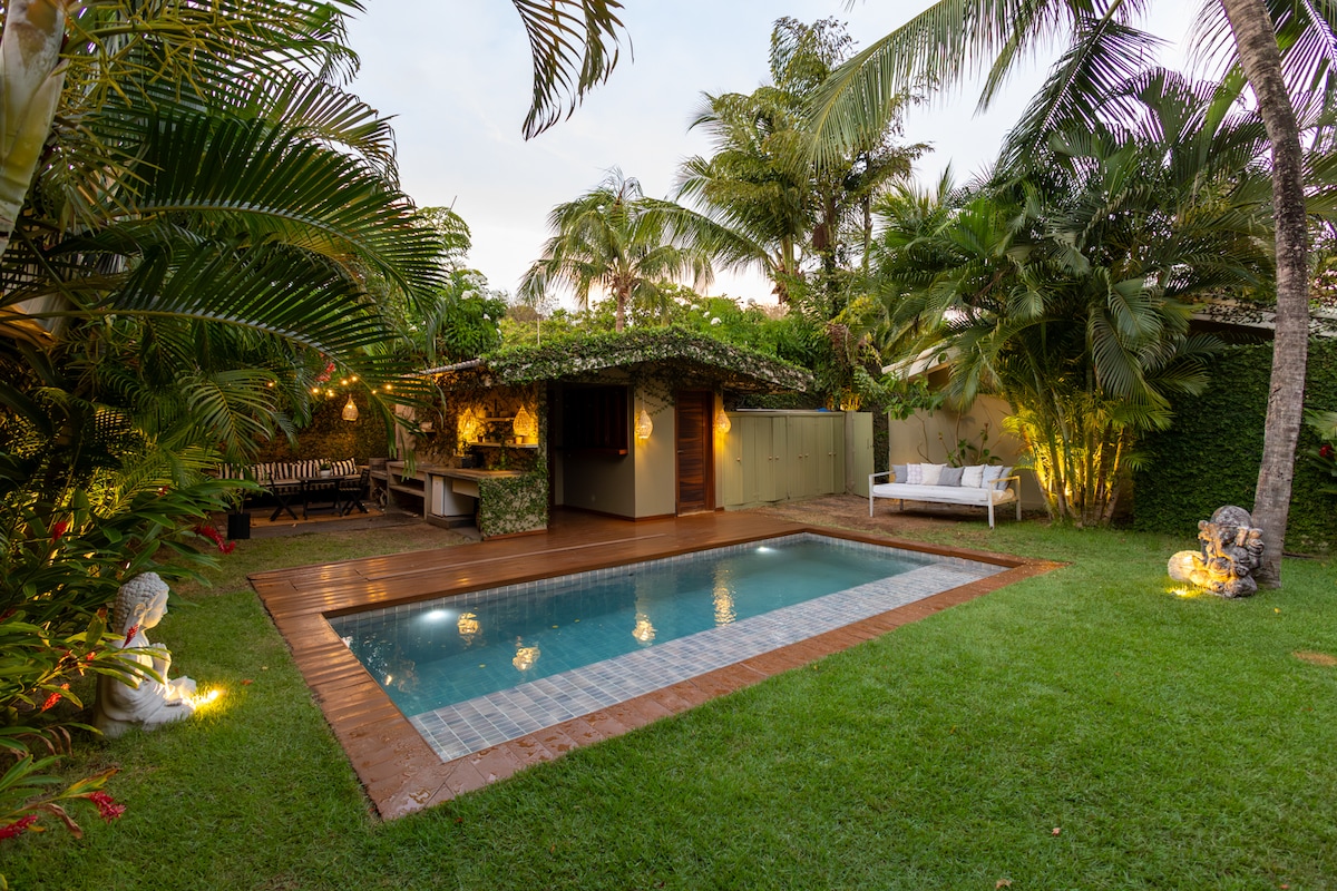 An inviting outdoor garden features a private pool surrounded by lush greenery. A wooden deck borders the pool, complemented by lounge seating and palm trees. Soft lighting enhances the tranquil atmosphere, encouraging relaxation in this serene environment.