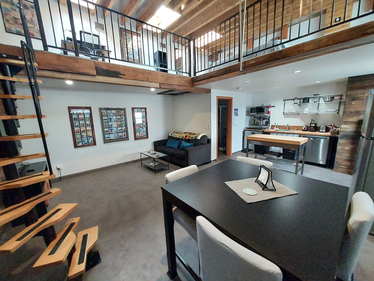 An open loft space is displayed with a spiral staircase leading to the upper level. The ground floor features a dining table, a cozy seating area, and a fully-equipped kitchen with modern appliances. Exposed beams and walls add character to the industrial-chic design.