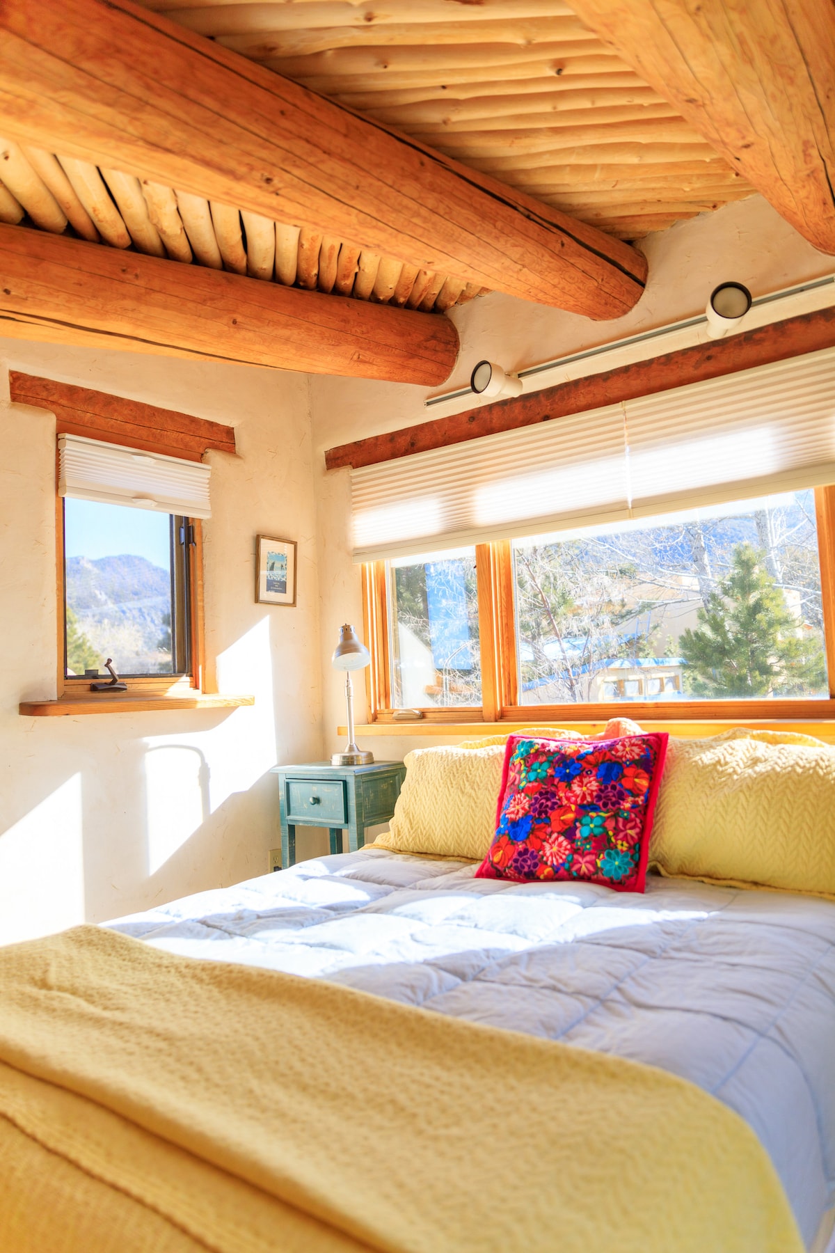 A cozy loft bedroom displays natural wooden beams overhead, with sunlight filtering through large windows. A colorful floral pillow rests on the spacious bed, alongside a soft yellow blanket. A bedside lamp and small table add functional touches to the inviting space.