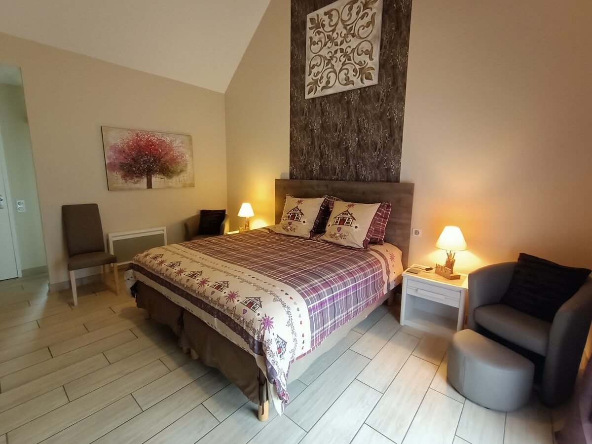 A spacious bedroom features a large bed with a patterned duvet and decorative pillows. Two bedside tables with lamps illuminate the room, complemented by a cozy seating area with two chairs. A piece of wall art adds a touch of color to the neutral walls.