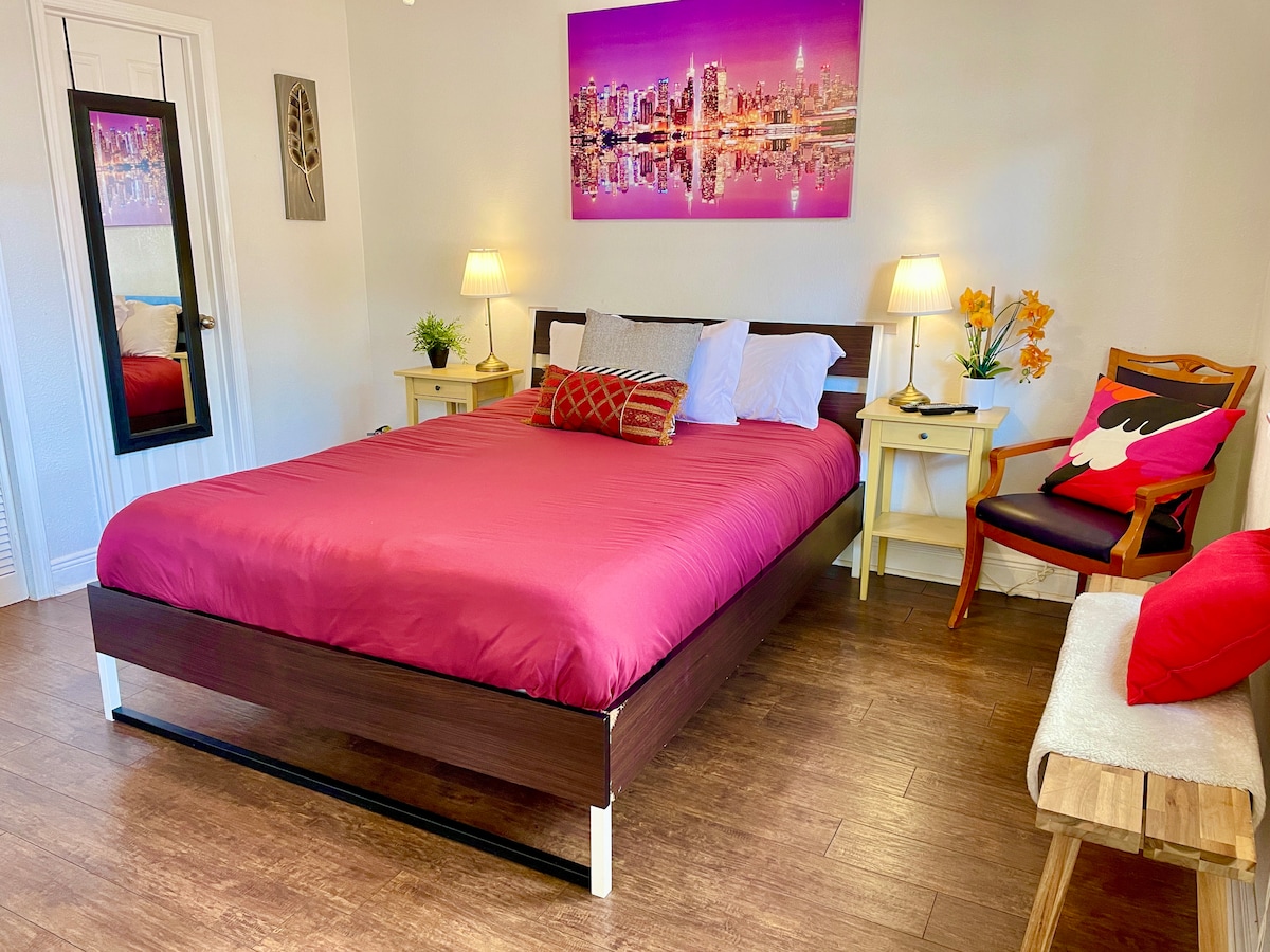A spacious sleeping area features a queen-size bed with a vibrant maroon coverlet and decorative pillows. Two nightstands with lamps are positioned on either side of the bed. A colorful cityscape artwork adorns the wall, while a plush chair and a small side table complete the cozy setup.