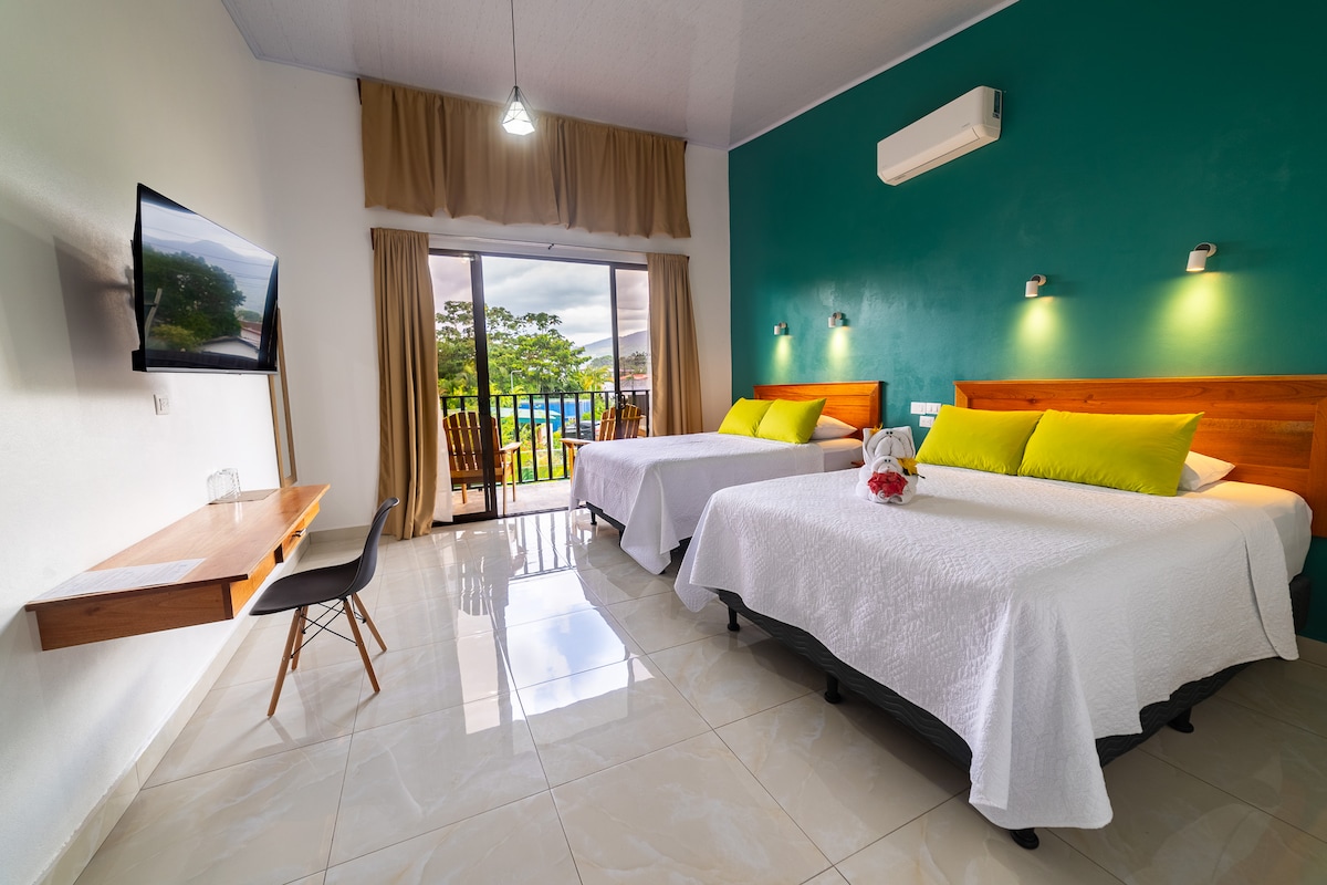 A bright room features two spacious beds adorned with white linens and vibrant green accent pillows. A flat-screen TV is mounted on the wall, and natural light floods in through large sliding glass doors, revealing a view of greenery outside. A small writing desk is positioned against the wall.