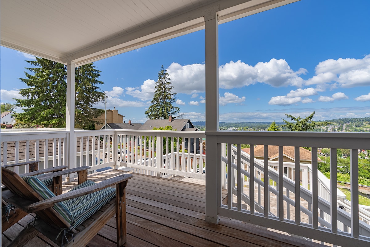 A spacious front deck is visible, featuring wooden flooring and two comfortable chairs with blue and green striped cushions. Panoramic views of lush greenery and distant hills are presented under a clear blue sky dotted with fluffy clouds.
