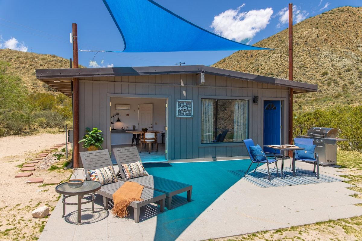 The cottage exterior features a welcoming blue front door and a shaded outdoor patio with comfortable lounge chairs. A small dining area and barbecue grill are visible, set against a backdrop of scenic desert hills and vibrant blue skies.