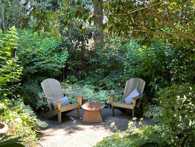 A serene patio area is surrounded by lush greenery, featuring two wooden Adirondack chairs facing a central fire pit. Various plants and foliage create a private and tranquil atmosphere, inviting relaxation in a natural setting.
