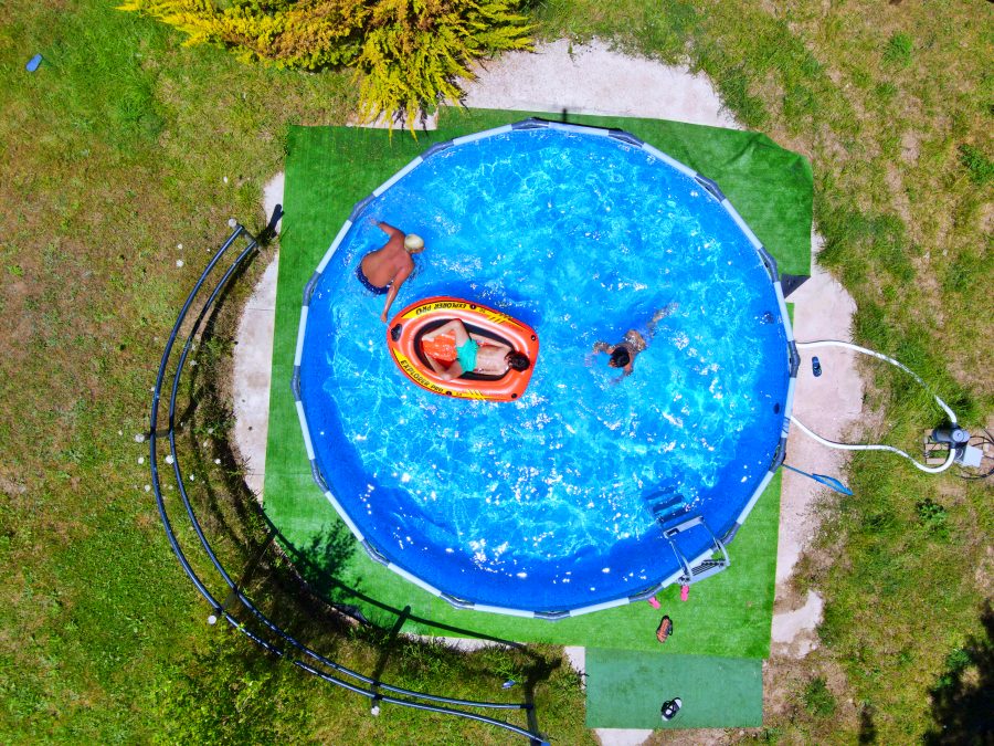 An aerial view of a circular pool is captured, showcasing vibrant blue water. A kayak is positioned in the pool, with two individuals enjoying the water. Surrounding the pool are patches of green grass and a small area of astroturf, enhancing the outdoor setting.