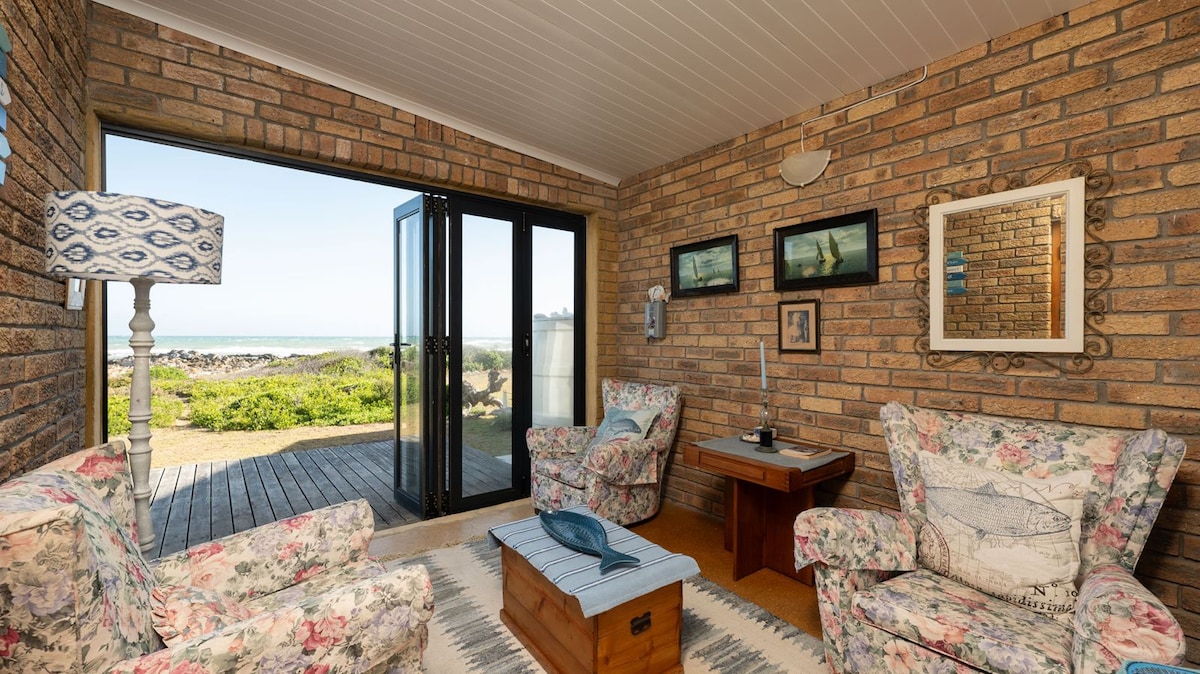 A cozy seating area features two floral armchairs and a wooden coffee table. Large glass doors open to a scenic outdoor view, allowing natural light to fill the room. A lamp stands elegantly in the corner, complementing the textured brick walls.